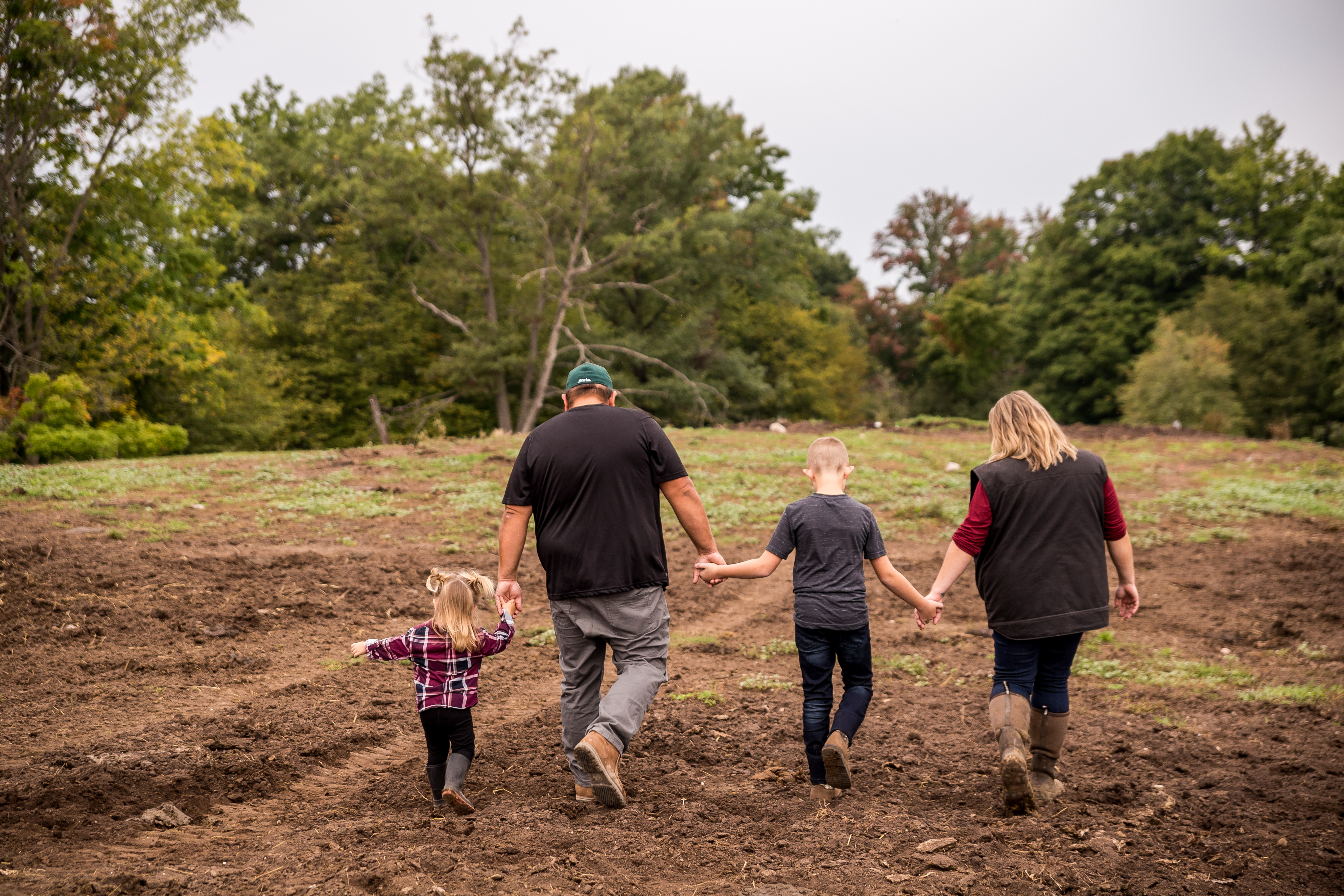 A man, a woman, and two children holding hands as they walk away from the camera through an unplanted field.