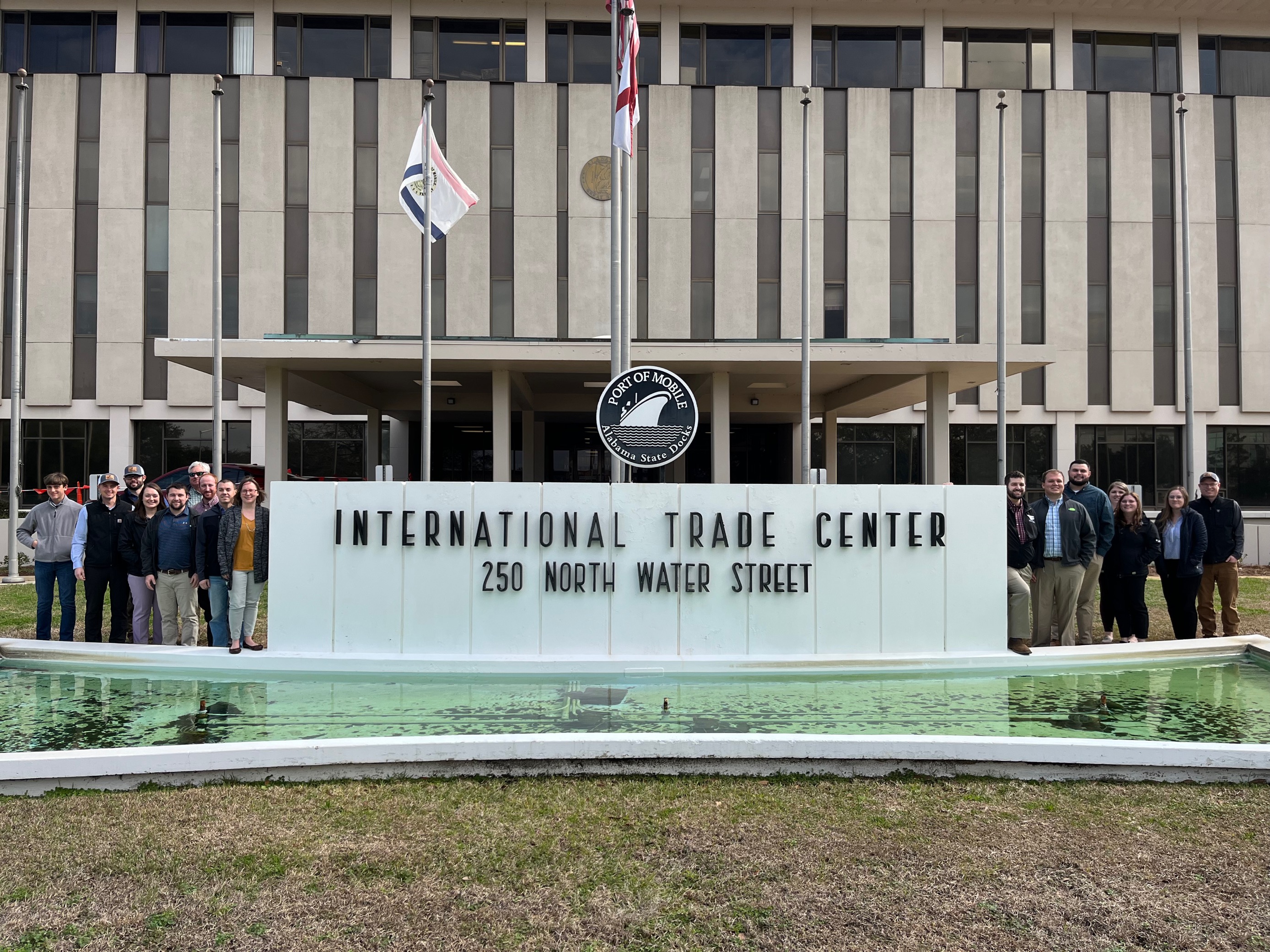A group of young farmers standing on either side of a sign reading "INTERNATIONAL TRADE CENTER" in front of a large government facility.