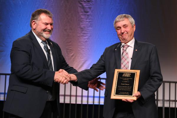 Presidential Volunteer of the Year award winner Hank Choate holding a plaque and shaking hands with MFB president Carl Bednarski while looking at the camera on stage at the 2022 MFB State Annual Meeting.
