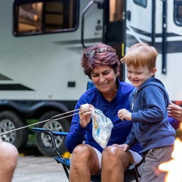 child and adult roast a marshmallow in front of a camper vehicle