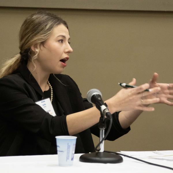 A young woman speaking into a microphone and gesturing to her left with both hands while she participates in a Young Farmer discussion meet.