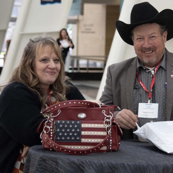 A man in a cowboy hat and a woman with an American flag-printed leather bag smiling at the camera while they stand in a large exhibit hall.